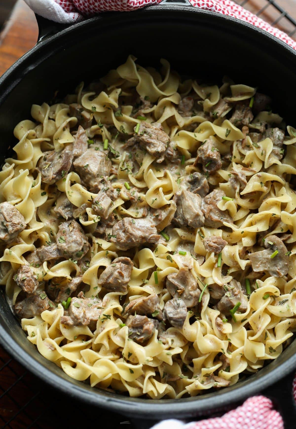 Overhead shot of a round, dark ceramic Crock Pot filled with egg noodles and chunks of beef in a creamy stroganoff sauce.