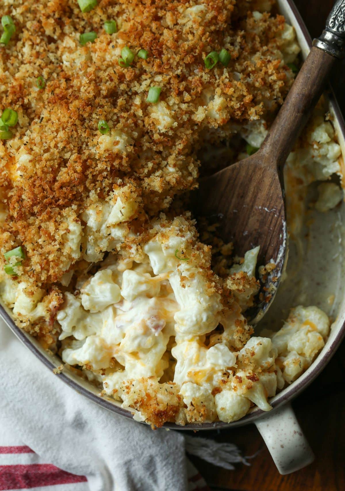 A cheesy cauliflower casserole with portions scooped out, and a wooden spoon resting in the dish.